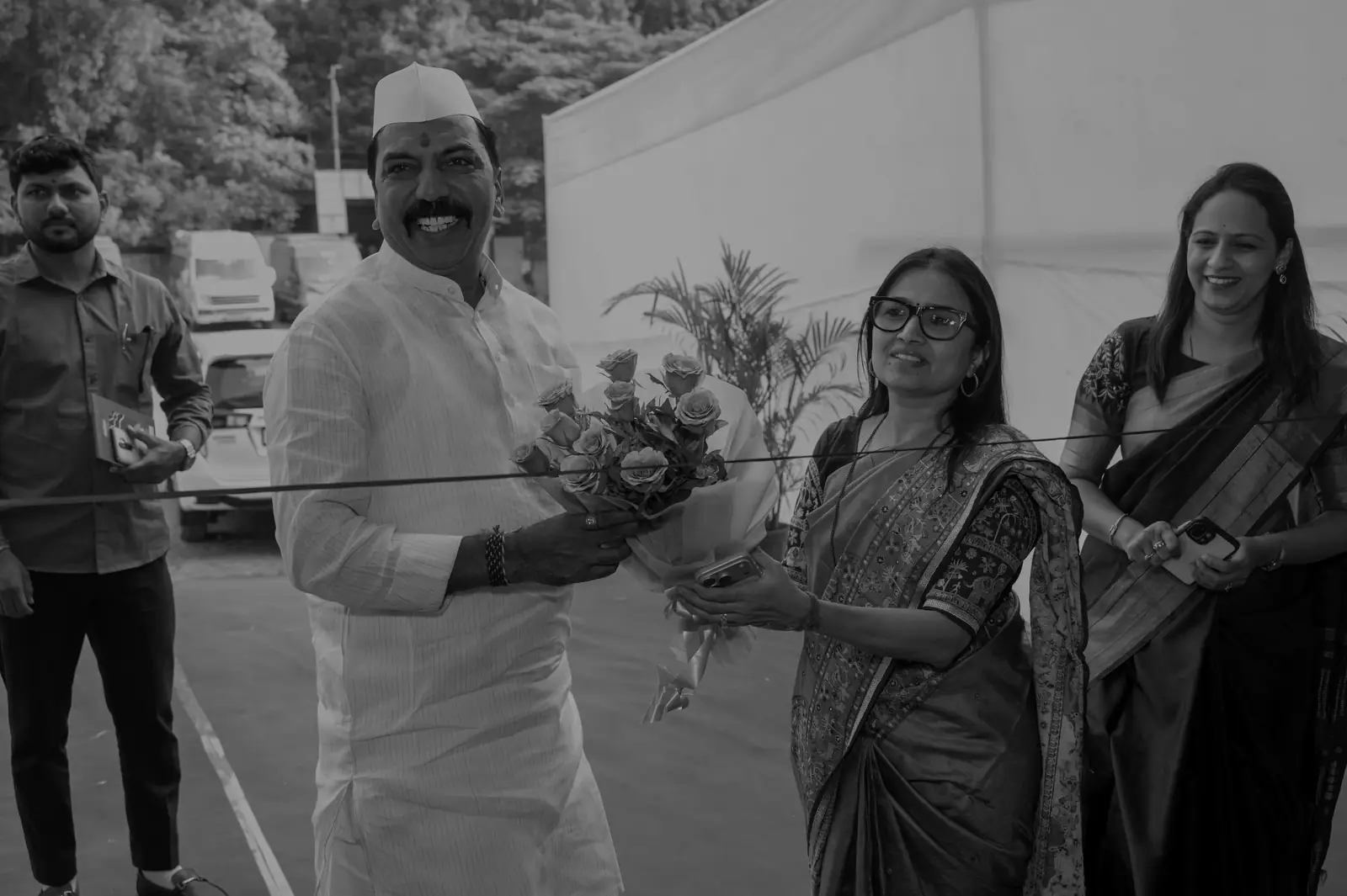 A man in a white kurta and cap holds a bouquet beside a red ribbon across the entrance of Gandhi Shilp Bazaar, with two women in saris smiling beside him