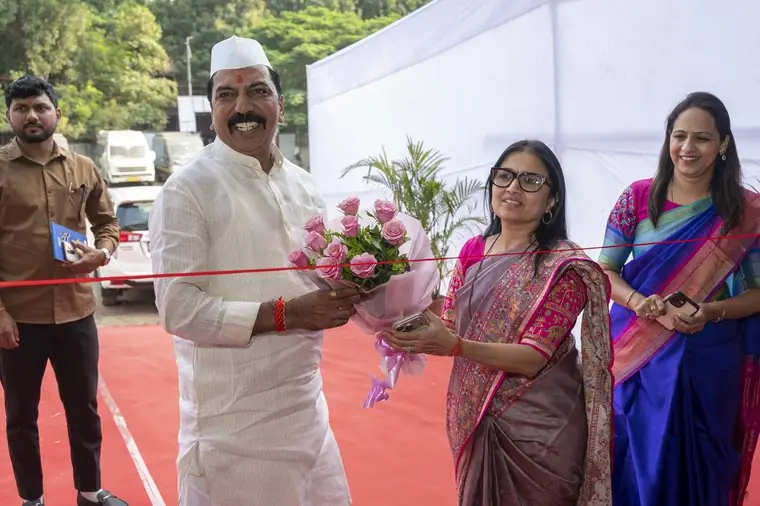 A man in a white kurta and cap holds a bouquet of pink roses beside a red ribbon stretched across the entrance, with two women in saris smiling beside him