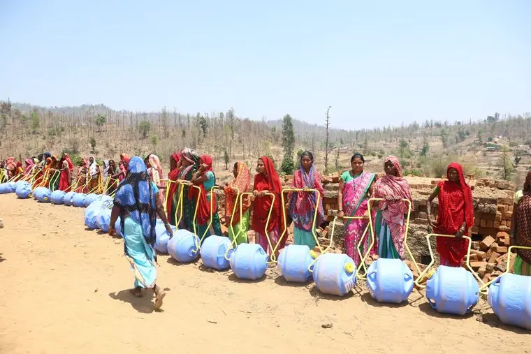 Women in red and pink saris stand in a line behind blue roller drums on dry ground, with hills behind